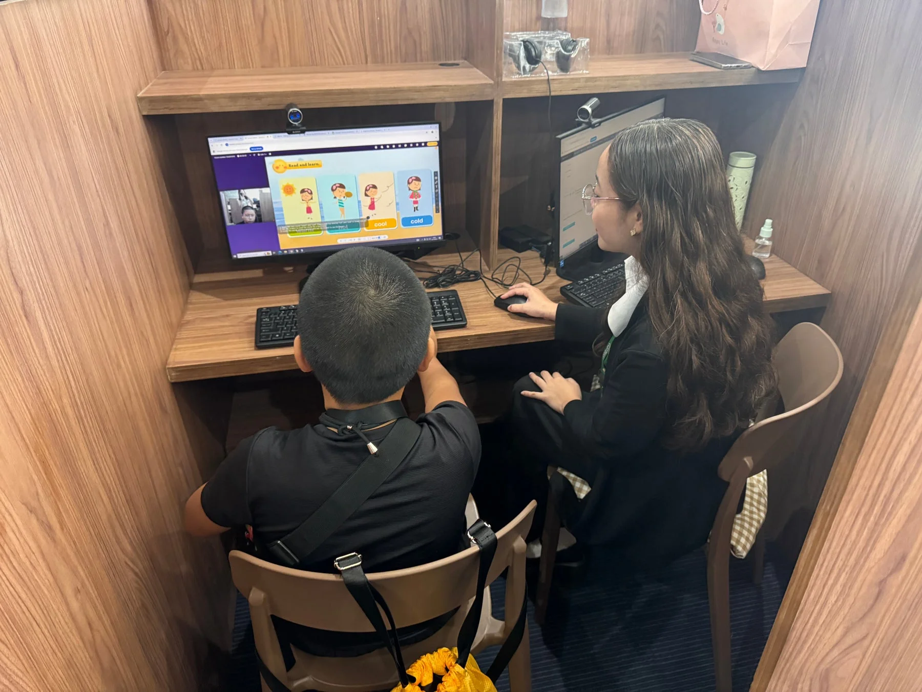 木製のブース内で、子供と大人が並んでパソコンを使用しています。画面には「Read and learn」と書かれた教材が表示されており、オンライン学習や個別指導の様子を捉えた一枚です。