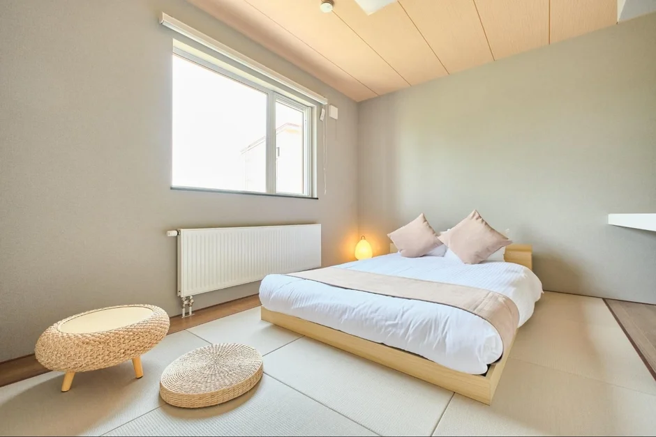 畳が敷かれた和モダンな寝室で、低い木製フレームのベッドが中央に置かれています。大きな窓から光が差し込み、シンプルで落ち着いた空間が広がっています。