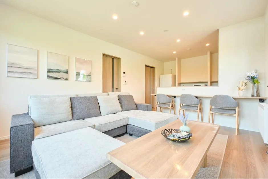 明るくモダンなリビングダイニングキッチンです。広々としたL字型ソファと木製コーヒーテーブルが配置され、奥にはカウンターキッチンとダイニングチェアが見えます。ニュートラルな色合いと間接照明がおしゃれな空間を演出しています。