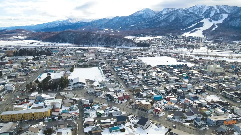 雪に覆われた冬の街並みを上空から捉えた一枚。遠景には雪化粧した山々が連なり、その一部にはスキー場のような白い斜面が見える。住宅や建物が広がり、寒々しいながらも美しい冬の都市景観が広がっている。
