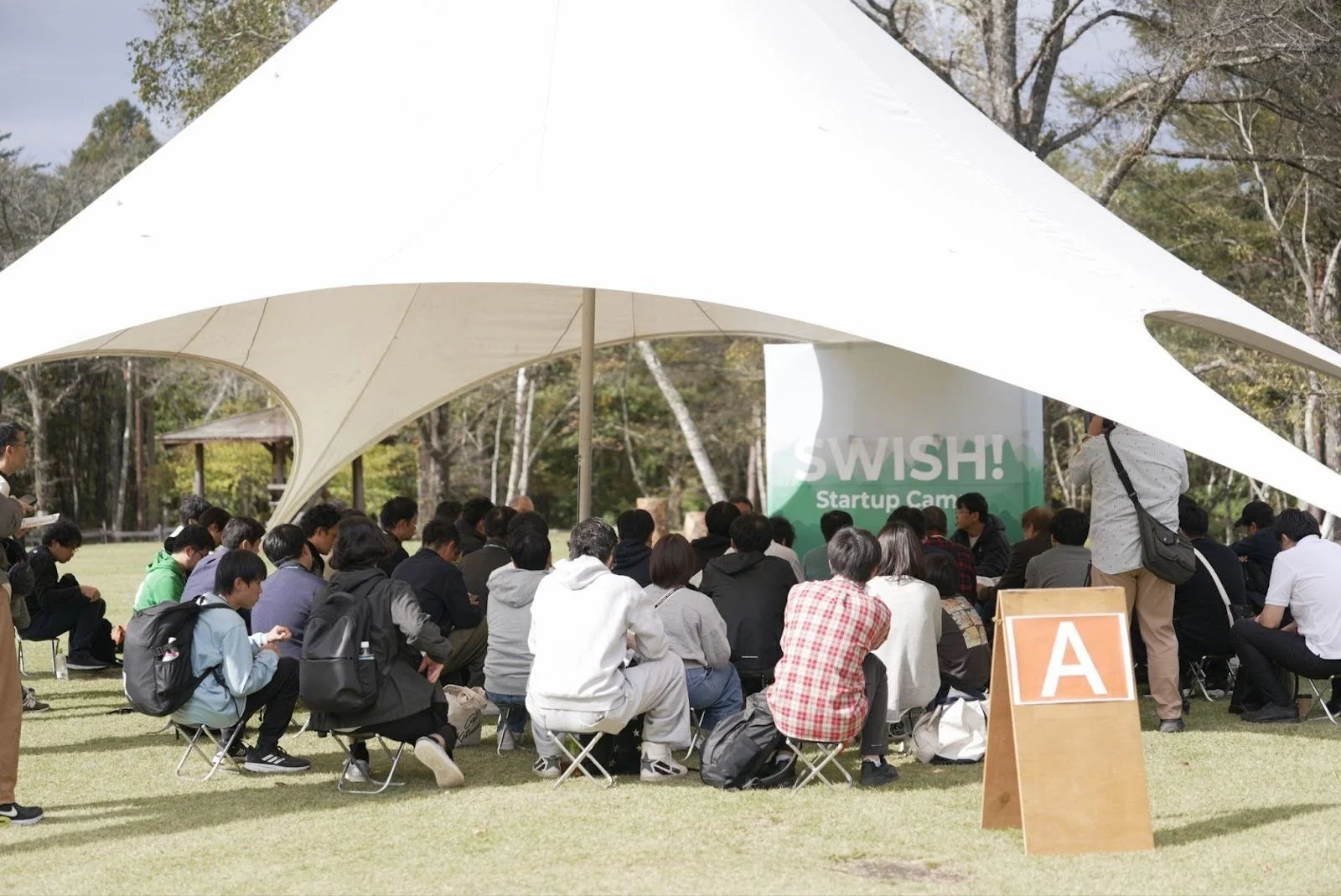 屋外イベントの様子、テントの下で話を聞く人々