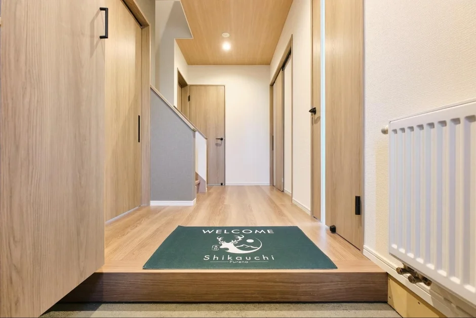 明るい木目調のフローリングと建具が特徴的な、モダンな玄関から廊下への眺めです。中央には「WELCOME Shikauchi Furano」と書かれた緑色のウェルカムマットが敷かれ、右奥には白いラジエーターが見えます。左手には階段があり、全体的に清潔感のある空間です。