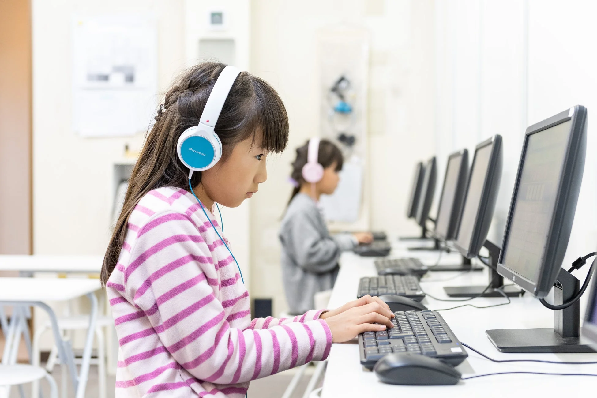 パソコン教室でヘッドホンを装着した子供たちが集中して学習している様子。手前の女の子がキーボードを操作しており、IT教育の場面を捉えている。
