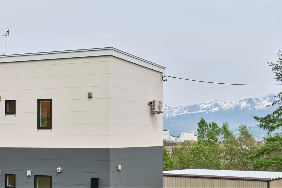 現代的なベージュとグレーのツートンカラーの建物が手前にあり、その背景には雪を頂いた壮大な山々が広がる風景です。