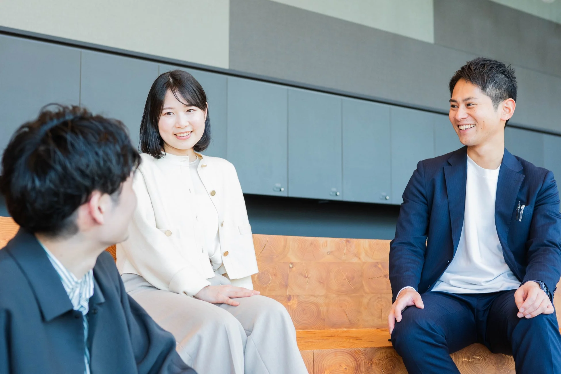 モダンな空間で、男女3人が木製のベンチに座り、笑顔で和やかに会話している様子