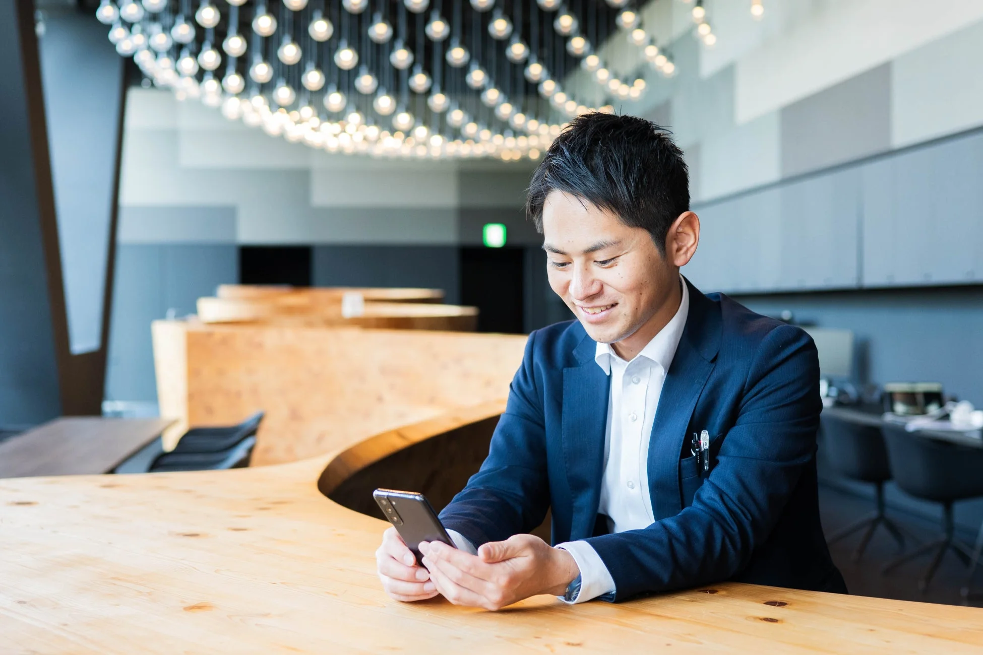 現代的なオフィスで、スーツを着た男性が木製のデスクに座り、スマートフォンを見ながら笑顔を見せている様子