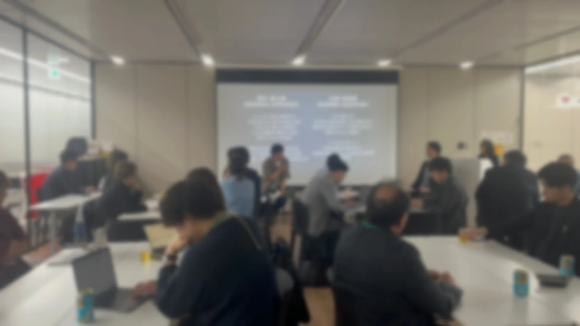 会議室のような空間で多くの人々が集まり、プロジェクターの画面を見たり、テーブルで作業したりしている様子