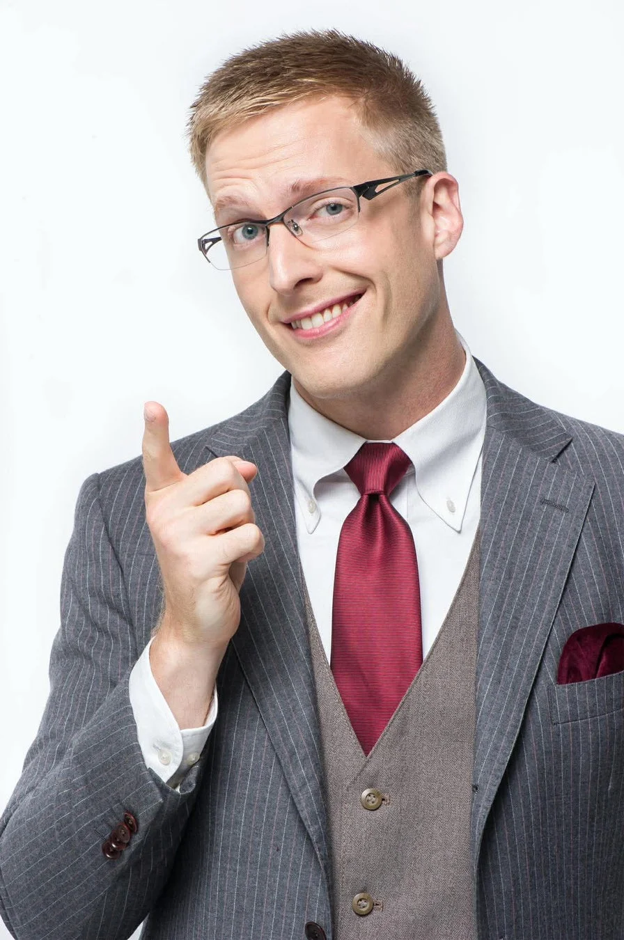 白い背景の前で、笑顔の男性がスーツと眼鏡を着用し、人差し指を立てて何かを指し示しているポートレートです。