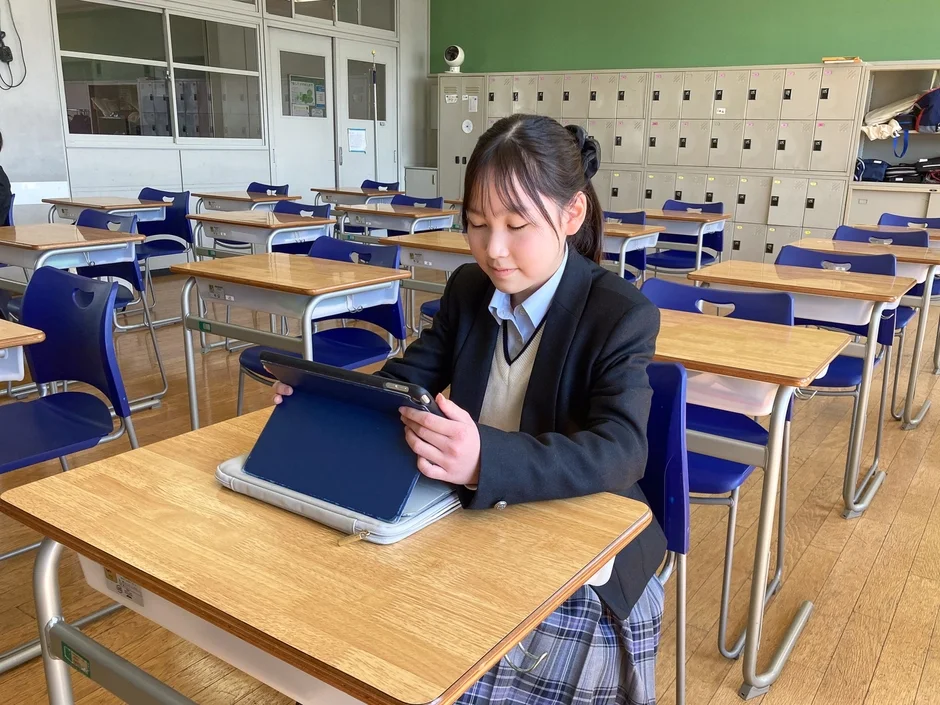 日本の学校の教室で、制服を着た女子生徒が机に置かれたタブレット端末を使用している