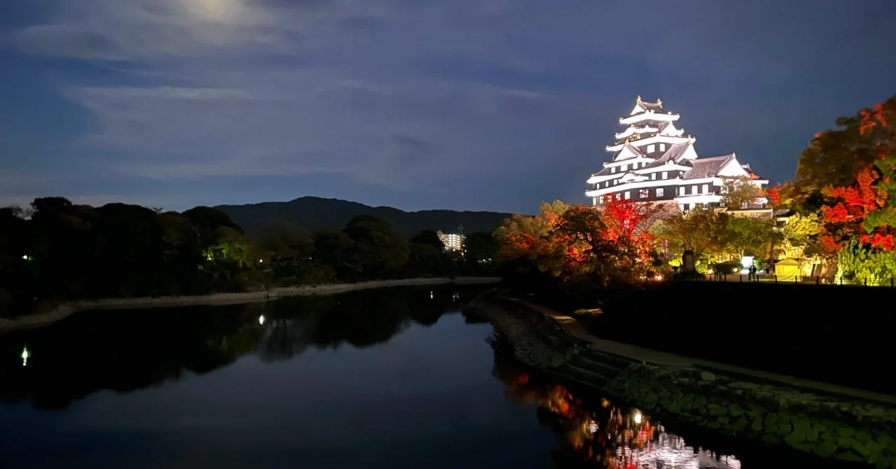 夜空の下、ライトアップされた日本の城が川面に美しく映り込んでいる。周囲の木々は紅葉し、赤や黄色の光で照らされ、幻想的な秋の夜景を演出している。