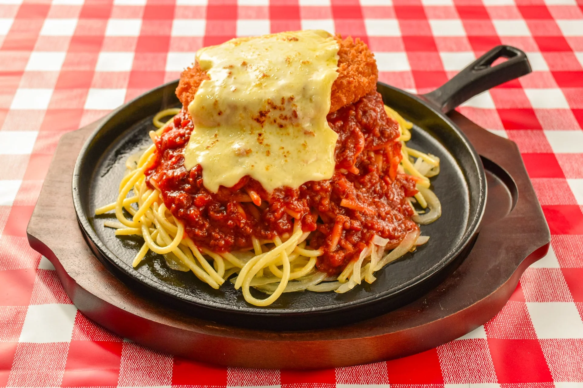 鉄板に盛られたスパゲッティの上に、たっぷりのミートソースと揚げたてのカツ、そしてとろけるチーズが乗った料理です。赤と白のチェック柄のテーブルクロスがレトロな雰囲気を醸し出しています。