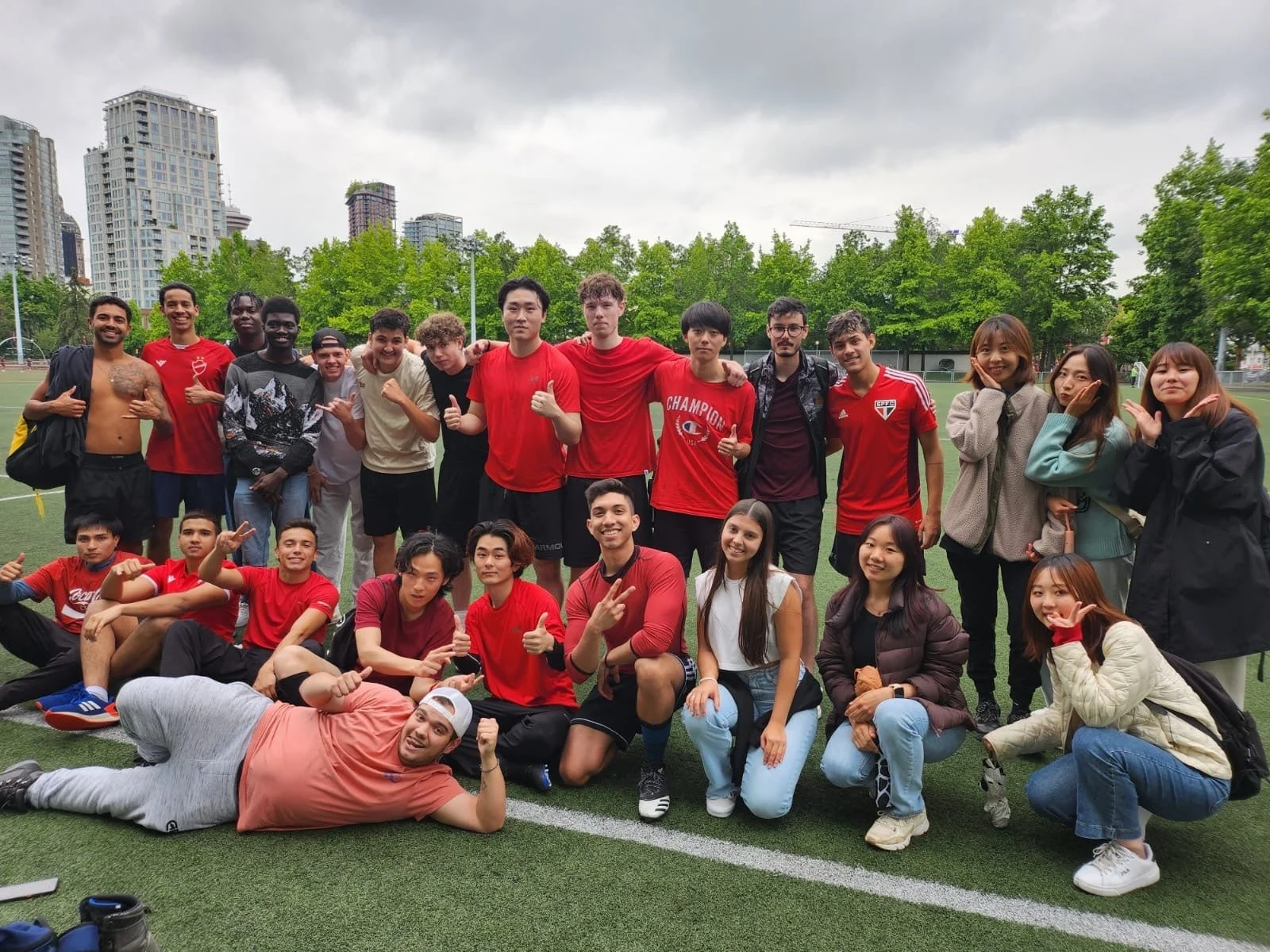 サッカー場のような緑のフィールドで、多くの若者が笑顔でポーズをとっている集合写真です。彼らの多くは赤いシャツを着用しており、背景には高層ビルや木々が見えます。和やかで楽しそうな雰囲気が伝わってきます。