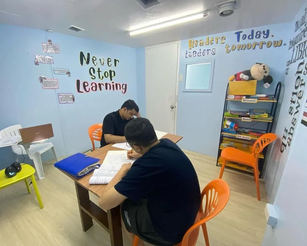 青い壁の部屋で、2人の男性が机に向かい勉強や作業をしている様子。