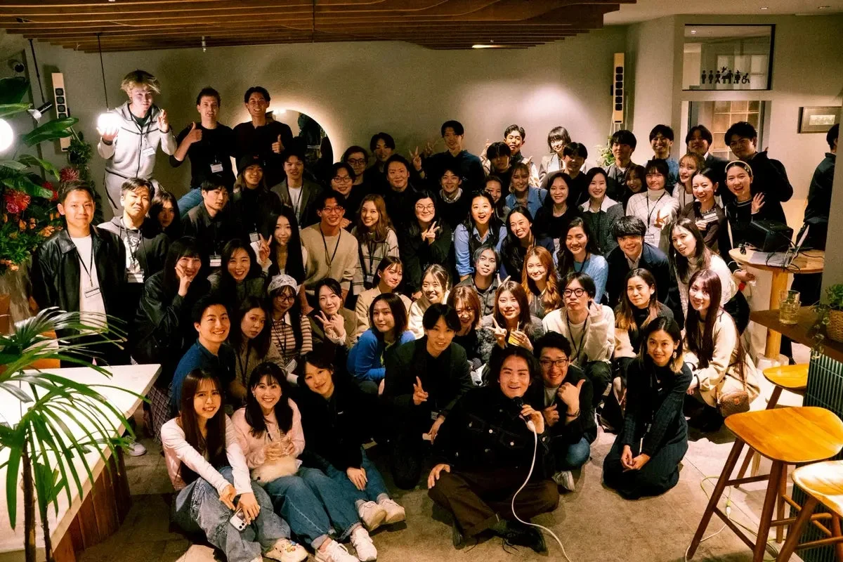 多くの若い男女が屋内で集合写真を撮っている様子。皆笑顔で、ピースサインをするなど、和やかな雰囲気で交流を楽しんでいる様子が伺えます。