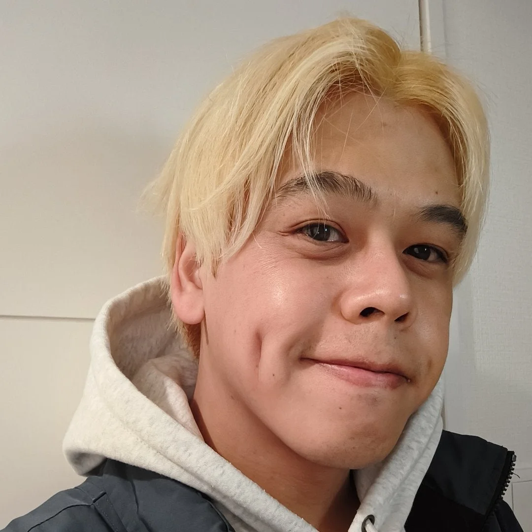 金髪の若い男性が白いパーカーとジャケットを着用し、カメラに向かって微笑んでいます