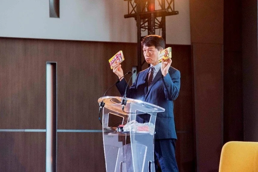 スーツを着た男性が透明な演壇に立ち、両手に小さな箱のような製品を持ってプレゼンテーションを行っています。会議やイベントでの発表風景と見られます。