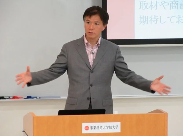 スーツを着た男性が講義室で両手を広げてプレゼンテーションを行っています