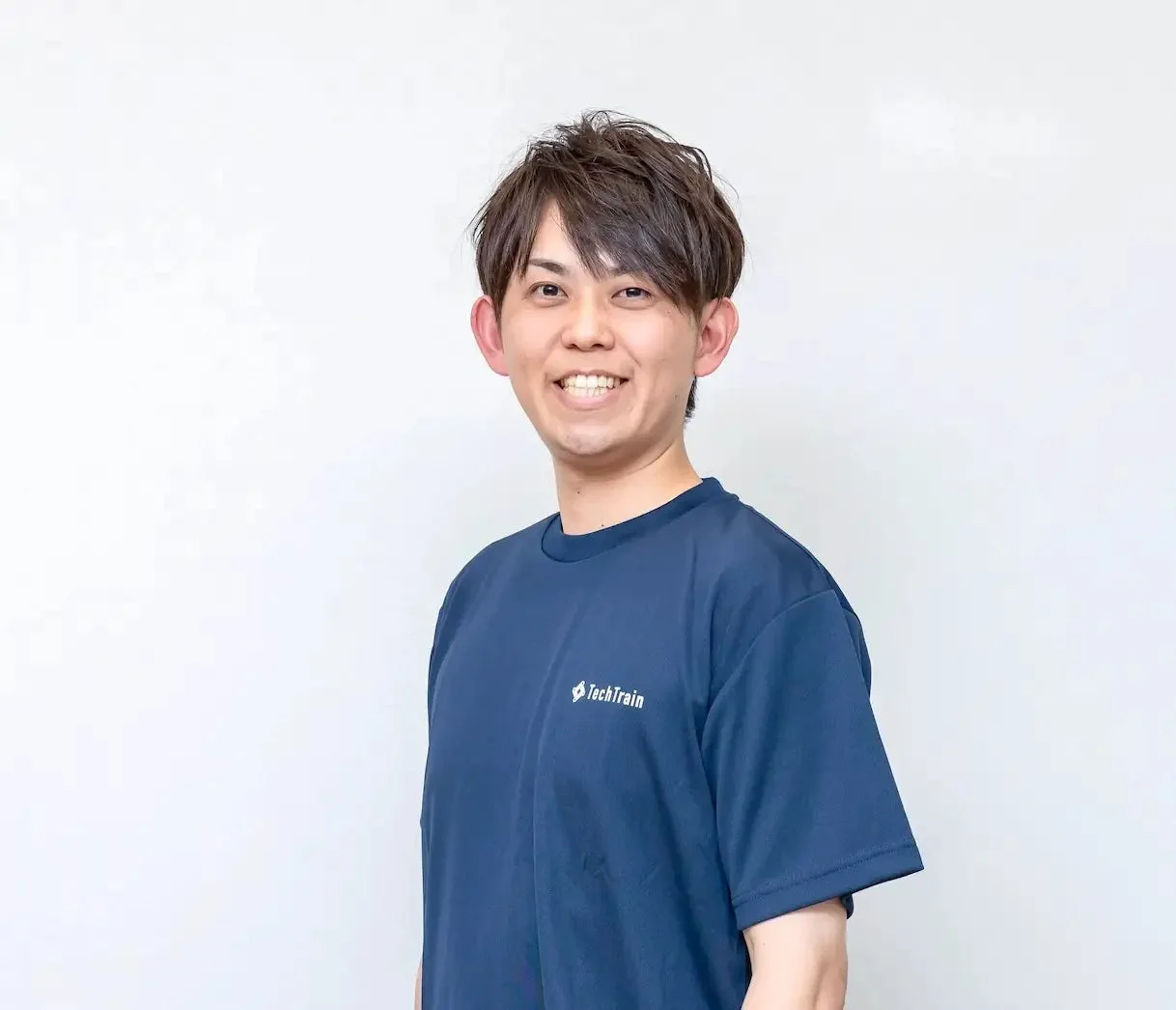 白い背景の前で紺色のTシャツを着た男性が笑顔でカメラを見ているポートレート写真