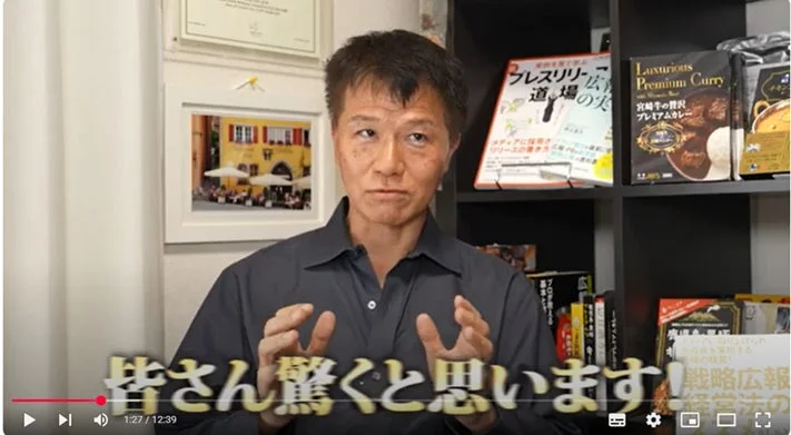 男性がカメラに向かって話しており、背景には本棚と商品が並んでいます。動画のテロップには「皆さん驚くと思います!」と表示されており、ビジネスや広報に関する内容のようです。