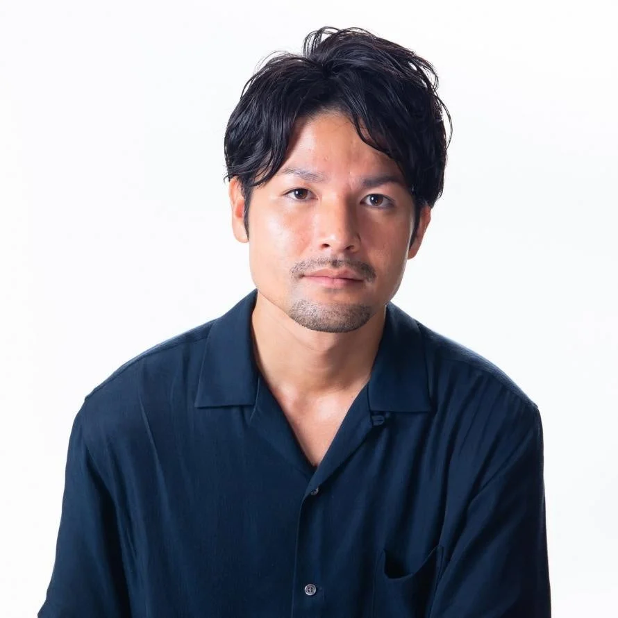 濃い色のシャツを着て、ひげを生やした男性のポートレート。白い背景の前でカメラをまっすぐ見つめています。