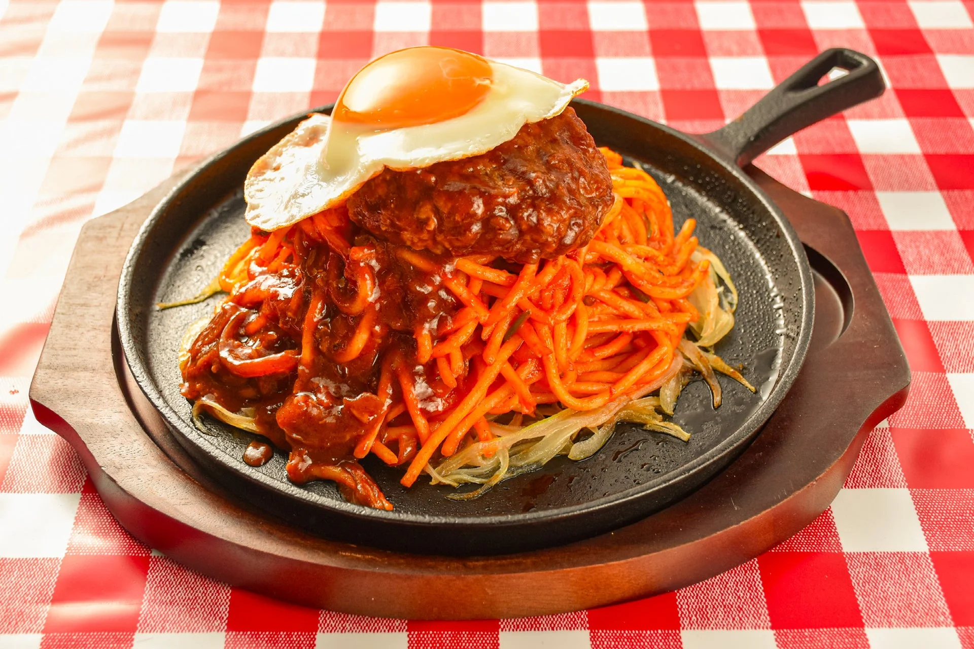 熱々の鉄板に盛られたハンバーグ、目玉焼き、そしてナポリタンが特徴的な洋食プレート。デミグラスソースがたっぷりかかり、赤いチェック柄のテーブルクロスがレトロな雰囲気を演出しています。食欲をそそる一品です。