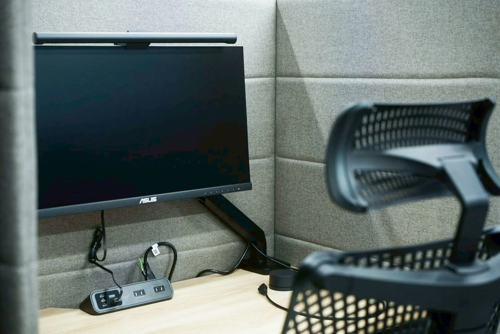 吸音材の壁に囲まれた集中できるワークスペースの様子です。ASUS製のモニター、モニターライト、電源タップが設置された木目調のデスクと、メッシュ素材のオフィスチェアが写っています。モダンで機能的な作業環境を示しています。
