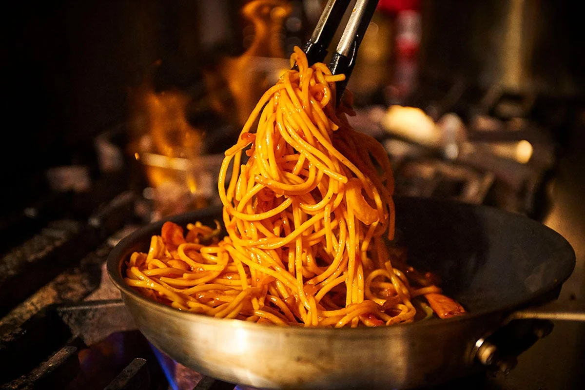 炎が上がるコンロの上で、トングを使って熱々のパスタまたは麺料理が炒められている調理風景。