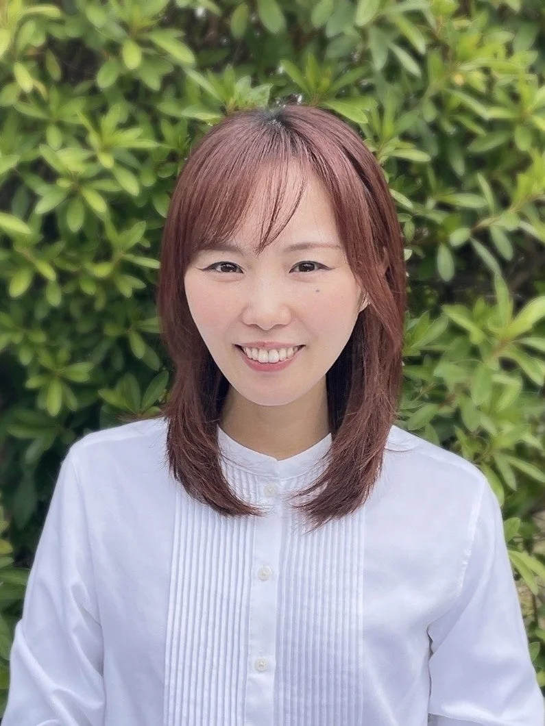緑の葉を背景に、白いシャツを着て笑顔でカメラを見つめるアジア人女性のポートレートです。髪はミディアム丈の茶色で、前髪があります。
