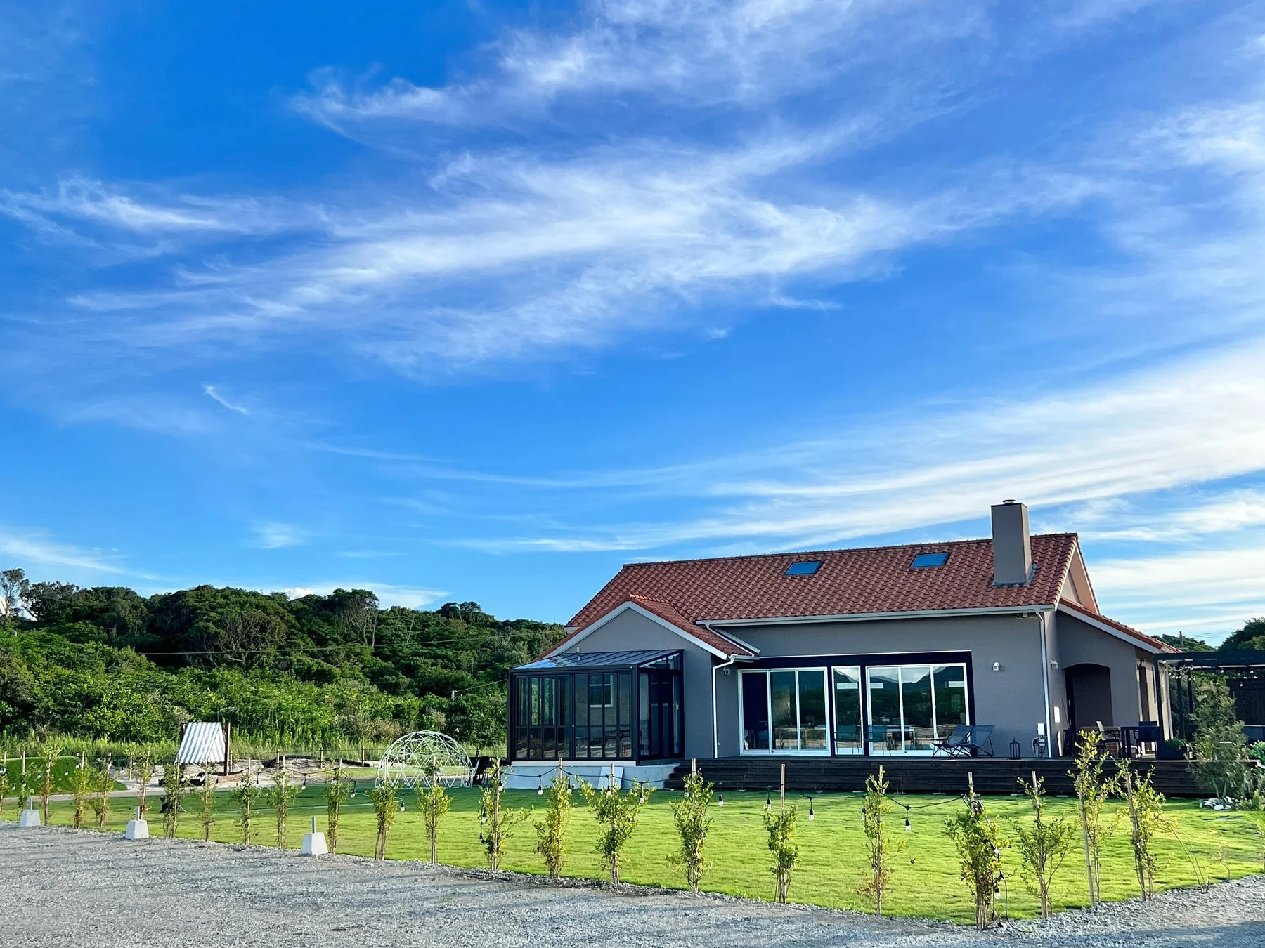 青い空と白い雲の下、赤い屋根とグレーの壁の家が広々とした庭に建っています
