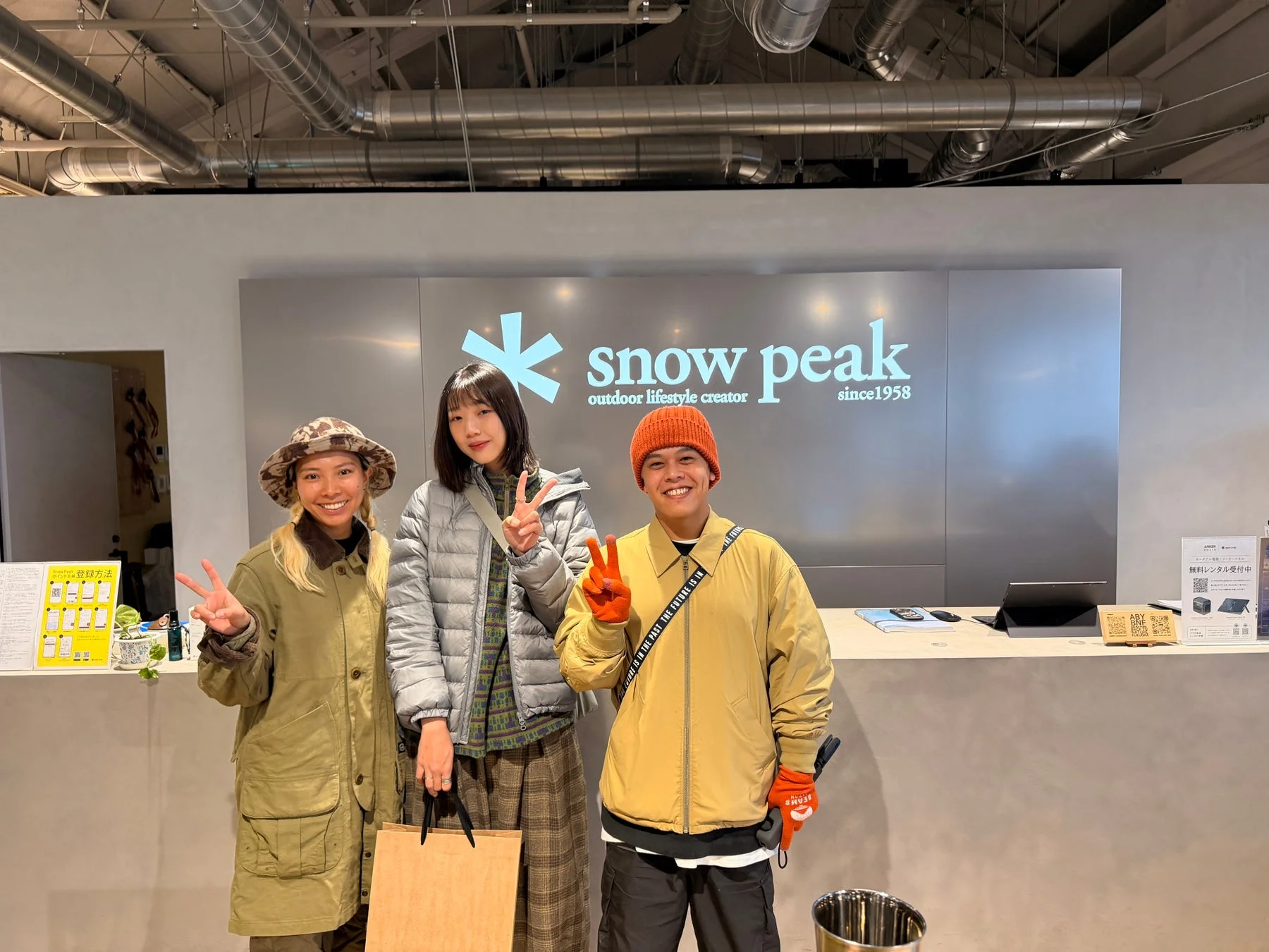 スノーピークの店舗内で、3人の男女が笑顔でピースサインをしながらポーズを取っています