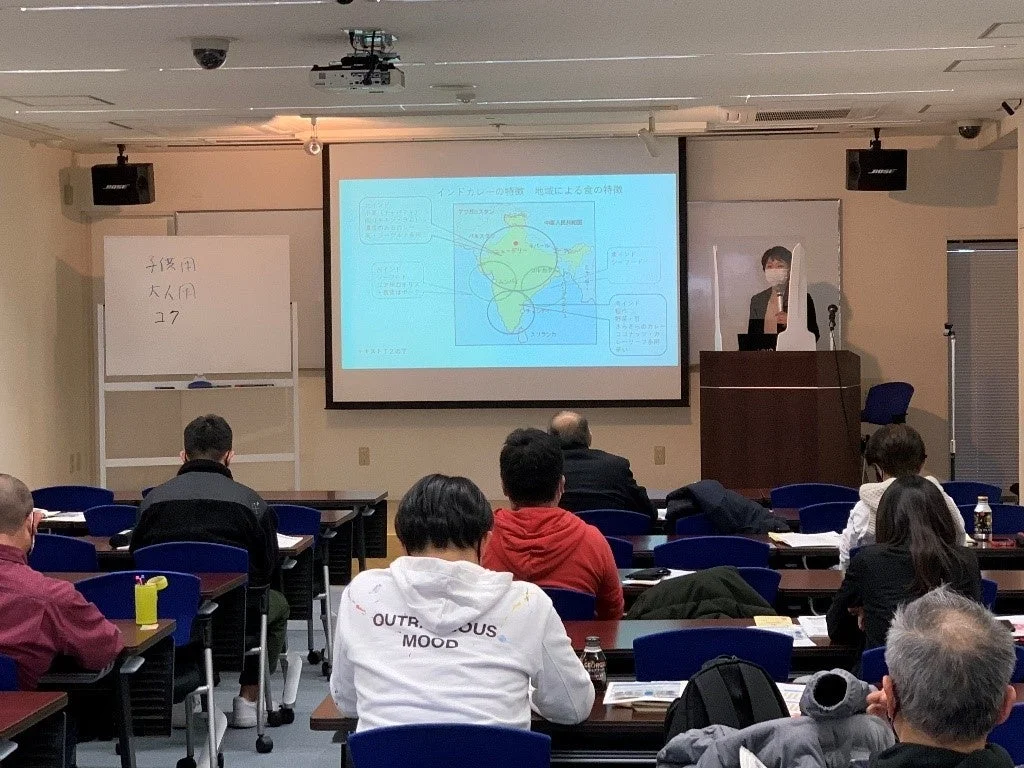 会議室で講師がインドカレーの地域特性についてプレゼンテーションを行っており、聴衆が熱心に耳を傾けている様子。スクリーンには地図や説明文が映し出され、ホワイトボードには日本語のメモが見える。