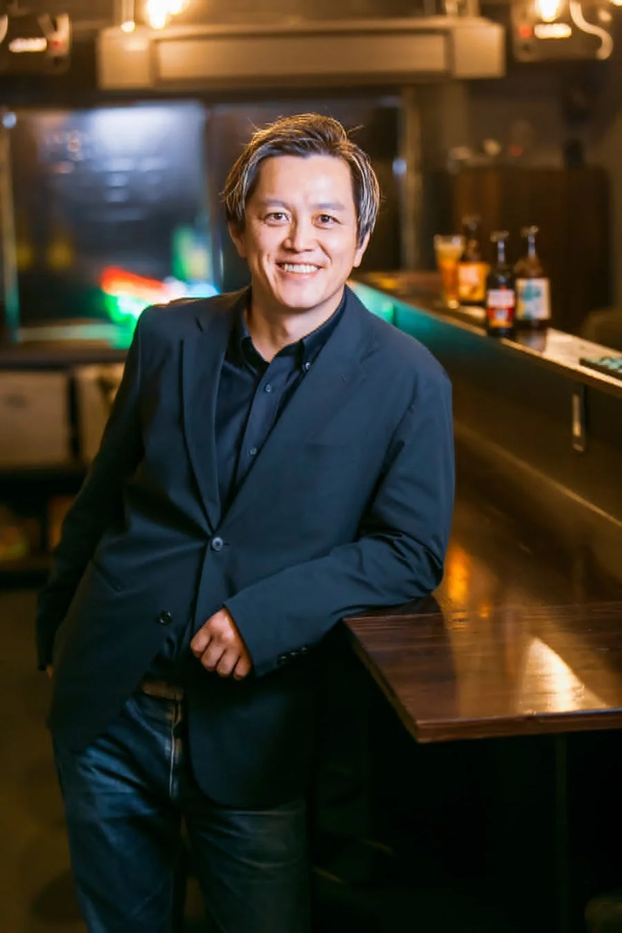 スーツジャケットとジーンズを着用したアジア人男性が、笑顔でバーのカウンターに寄りかかっています。背景にはボトルが並び、落ち着いた雰囲気のバーの様子が伺えます。