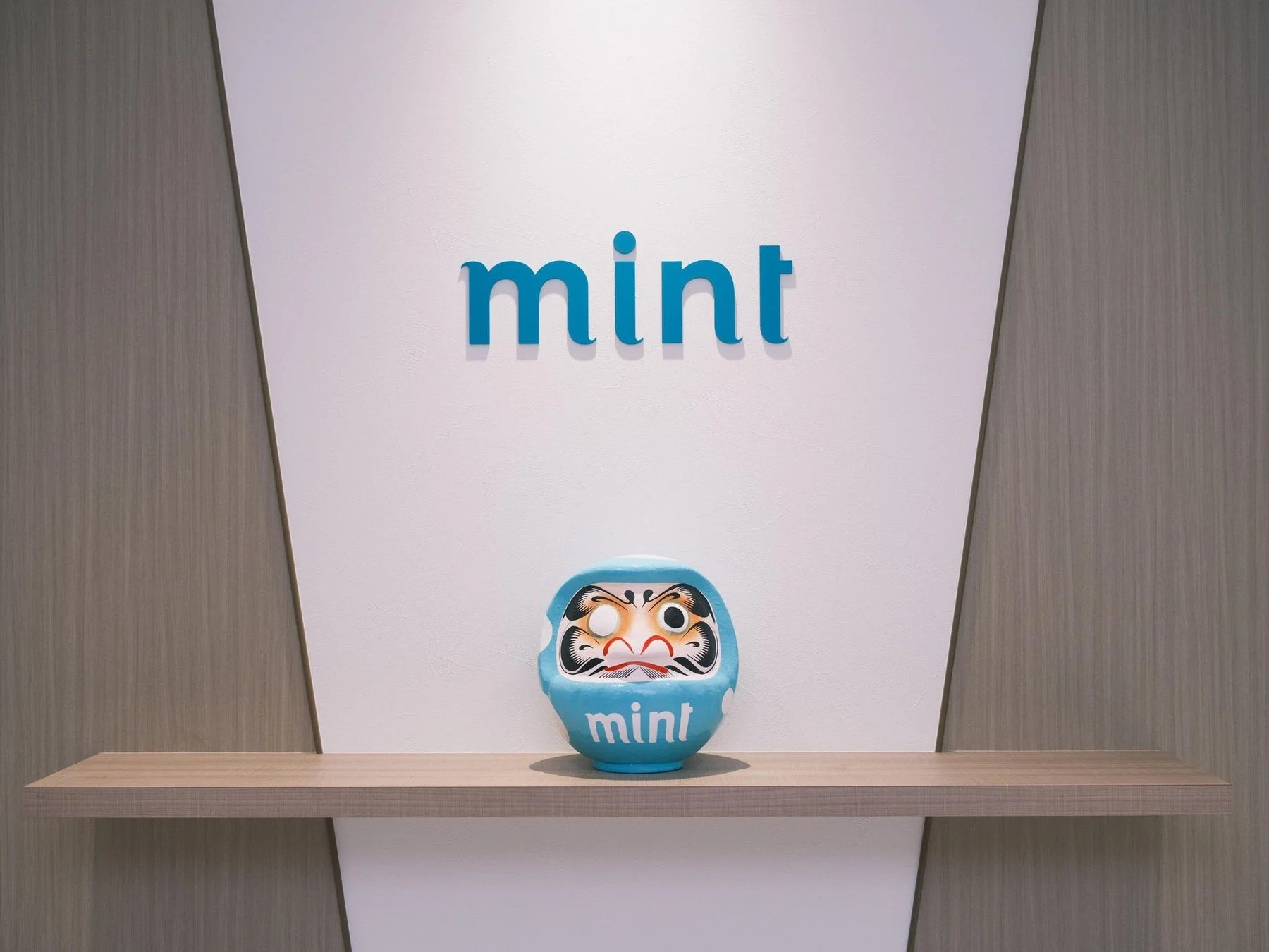 「mint」というロゴが大きく表示された白い壁の前に、青い達磨が木製の棚に置かれている様子