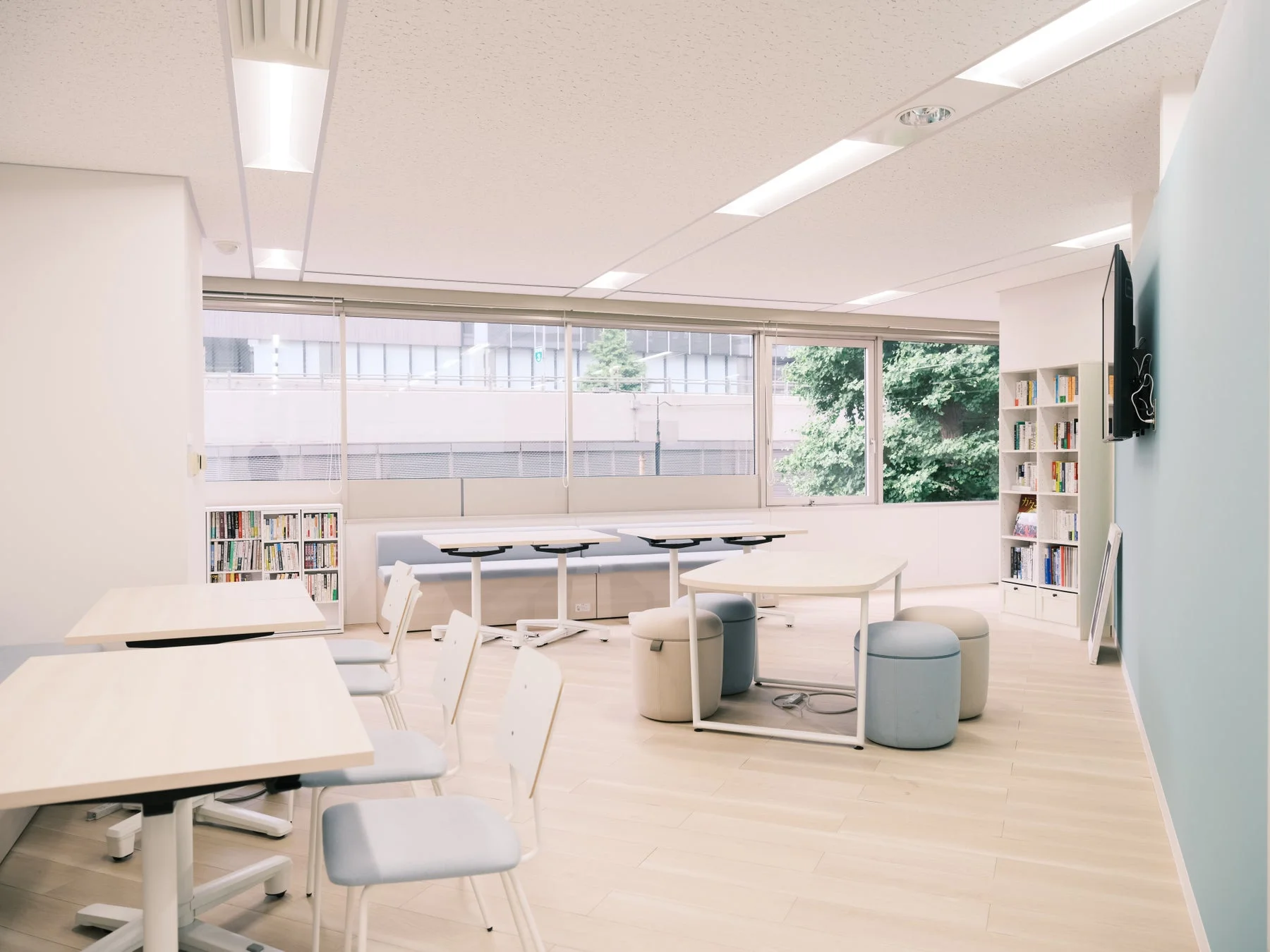 明るくモダンな雰囲気の室内空間。木目調の床に、淡い色のテーブル、椅子、クッションが配置され、大きな窓からは外の景色が見えます。