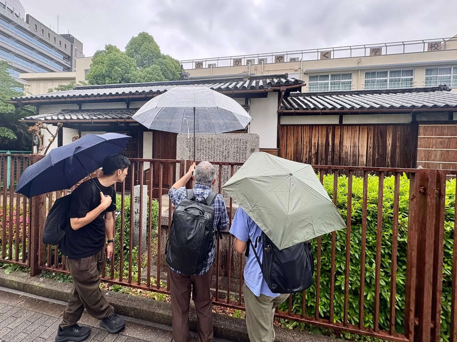 雨の中、傘をさした3人が瓦屋根の伝統的な日本家屋と石碑の前を歩いている