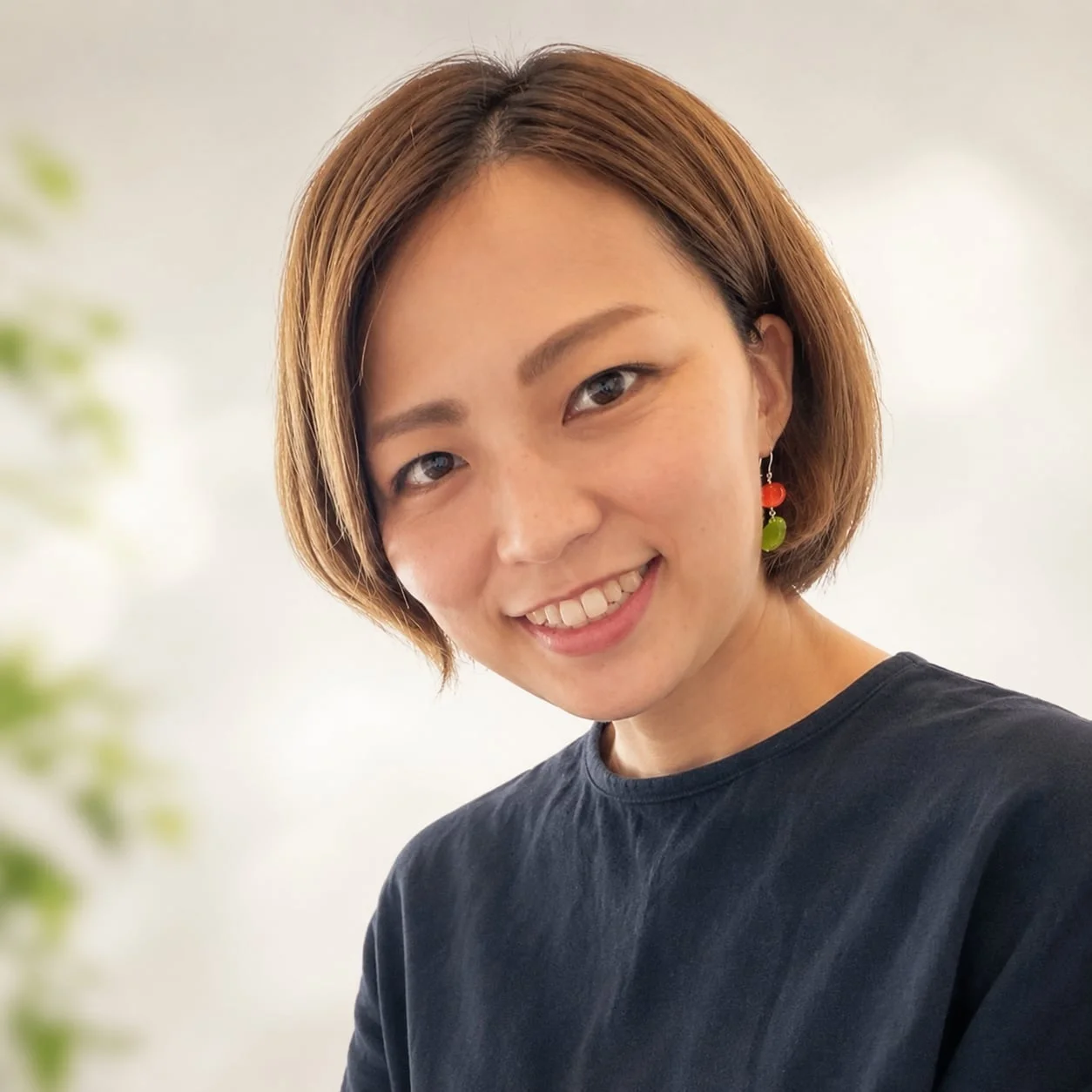 ショートヘアの女性が笑顔でこちらを見ているポートレート写真です。赤い玉と緑の玉のイヤリングを着用しており、明るく親しみやすい雰囲気です。