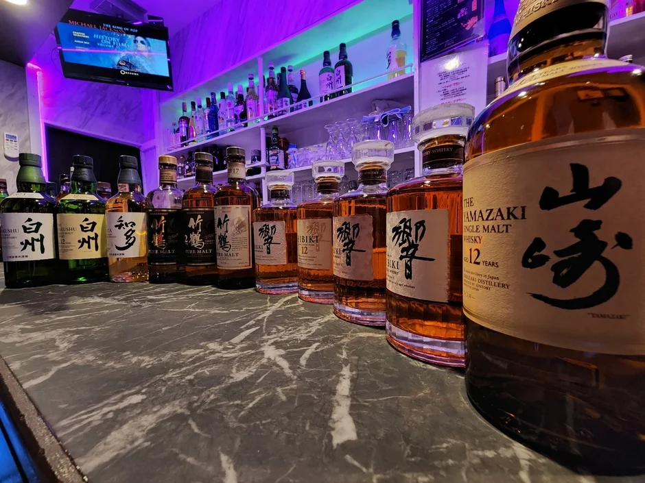 紫色の照明が印象的なバーカウンターに、山崎、白州、響などの様々なジャパニーズウイスキーのボトルがずらりと並んでいます。背景のテレビにはマイケル・ジャクソンの映像が映し出されており、おしゃれな空間が演出されています。