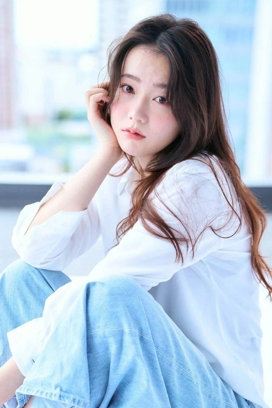 白いシャツとデニムを着用した若い女性が、柔らかな光の中で座ってカメラを見つめています。優しい表情が印象的なポートレート写真です。