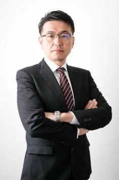 スーツと眼鏡を着用した中年の男性が、腕を組み真剣な表情でカメラを見つめているポートレート写真です。ビジネスシーンに適したフォーマルな印象を与えます。