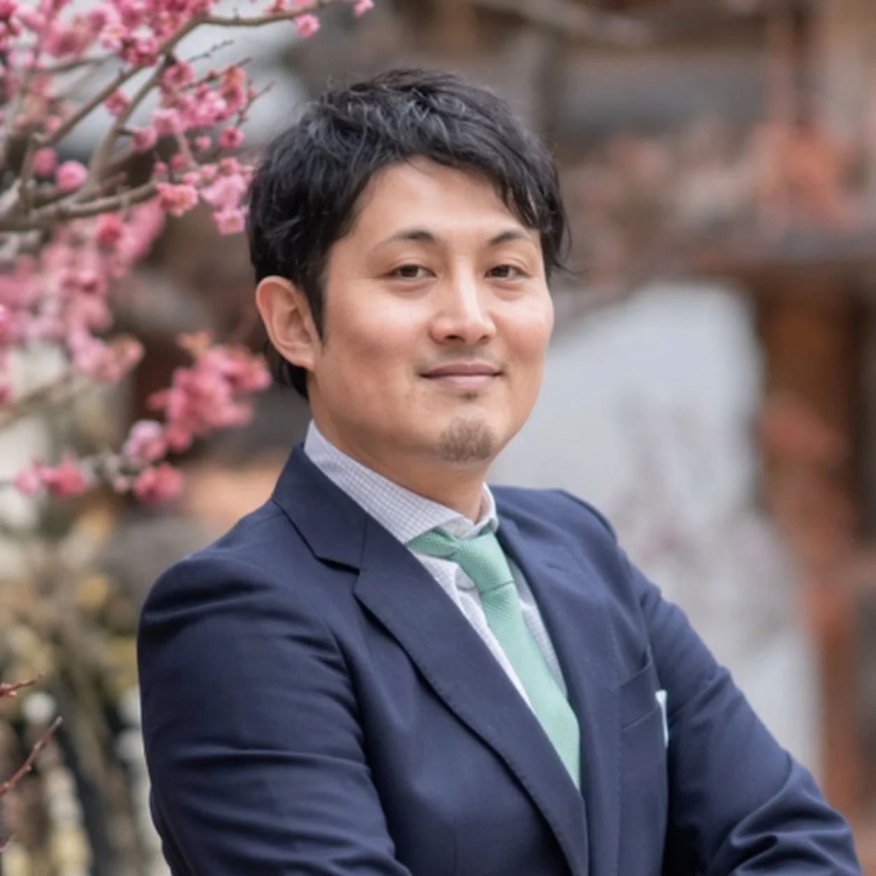 スーツ姿の男性が、背景の満開のピンク色の梅の花を背に、穏やかな笑顔を見せています。春の訪れを感じさせる明るい雰囲気の写真です。