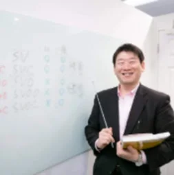 笑顔の男性教師がホワイトボードの前に立ち、ポインターと本を持って授業をしている様子です。ホワイトボードには英語の文法構造が書かれています。