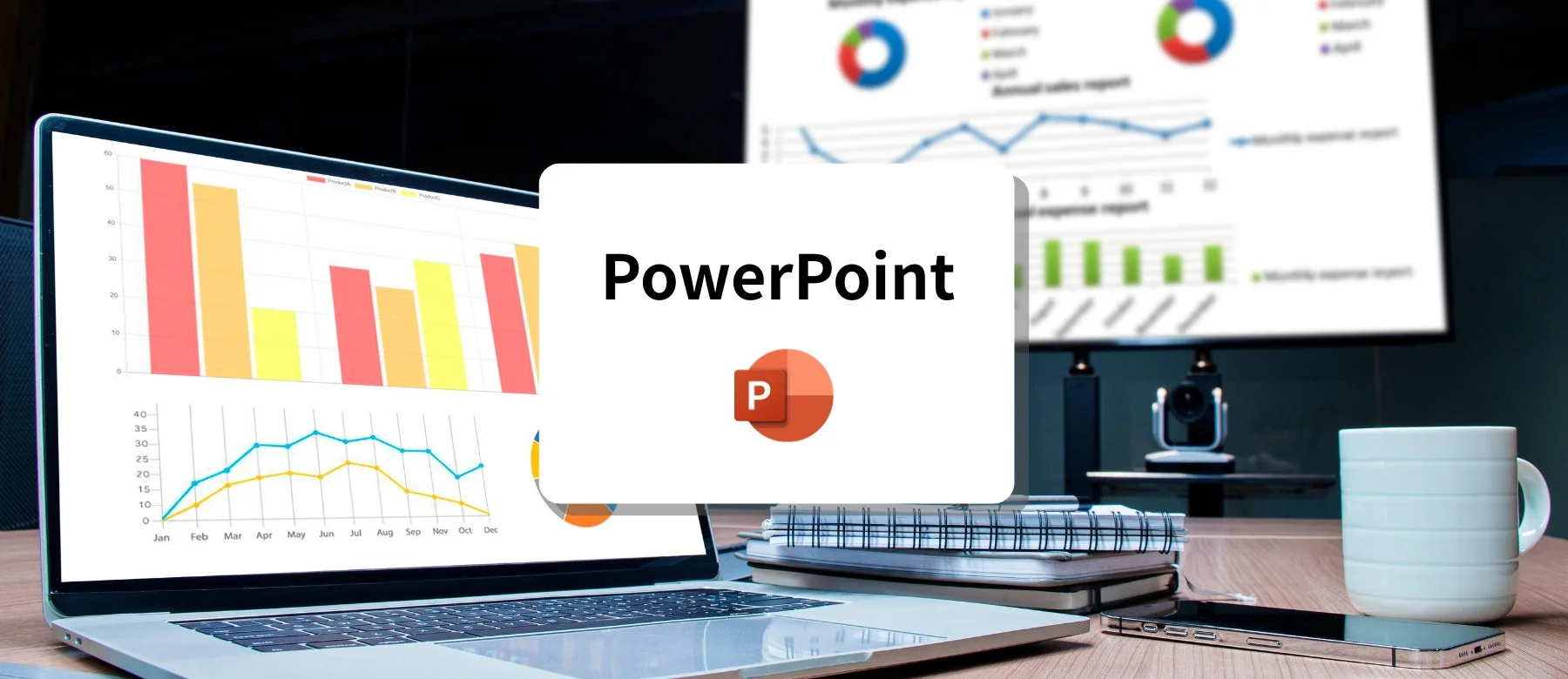 ノートパソコンと外部モニターにビジネスレポートのグラフが表示されており、手前にはPowerPointのロゴと文字が見えます。会議室のような空間で、資料作成やデータ分析が行われている様子を表しています。