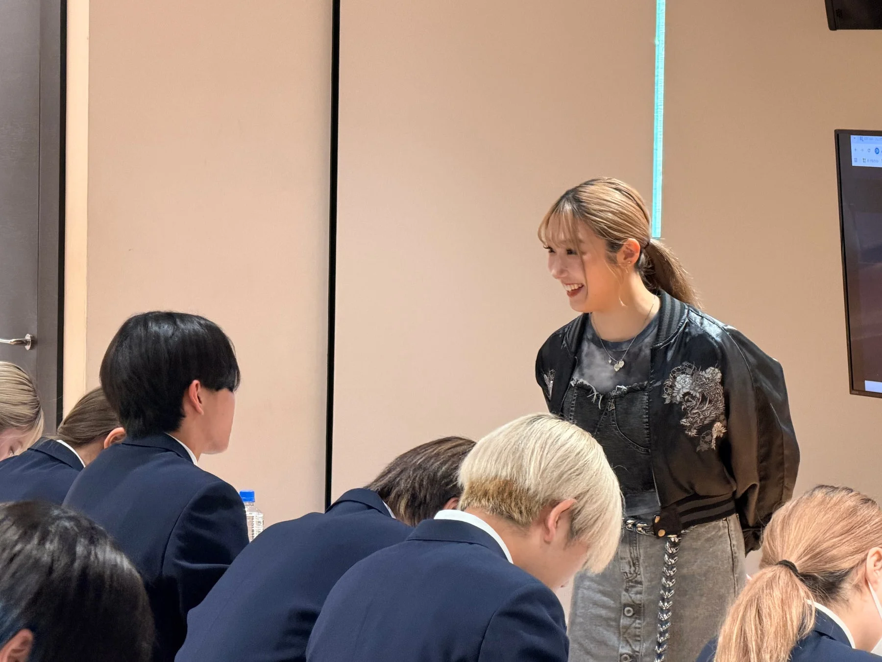 ボンバージャケットを着た女性が、制服姿の学生らしき人々に向かって笑顔で話している様子