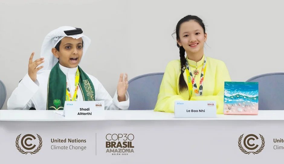 気候変動に関する国際会議「COP30」に参加していると思われる2人の若い子供が写っています。少年はアラブの伝統衣装を着用し熱心に話しており、少女は笑顔で着席しています。テーブルにはそれぞれの名前と、国連やCOP30のロゴが見えます。