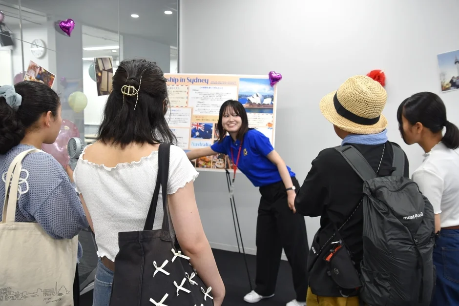 複数の学生がプレゼンテーションボードの前に集まり、青いシャツを着た女性がシドニーでのインターンシップについて説明しています