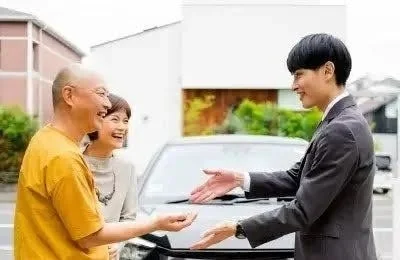 笑顔の夫婦が営業担当者と車の鍵らしきものをやり取りしている場面です。新車購入やレンタカーの受け渡しなど、車に関する契約の瞬間を表しています。