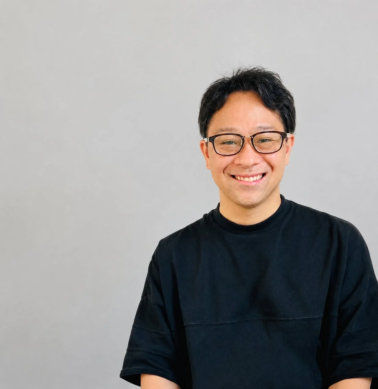 眼鏡をかけたアジア人男性が黒いTシャツを着て、シンプルな灰色の背景の前で笑顔を見せているポートレート写真