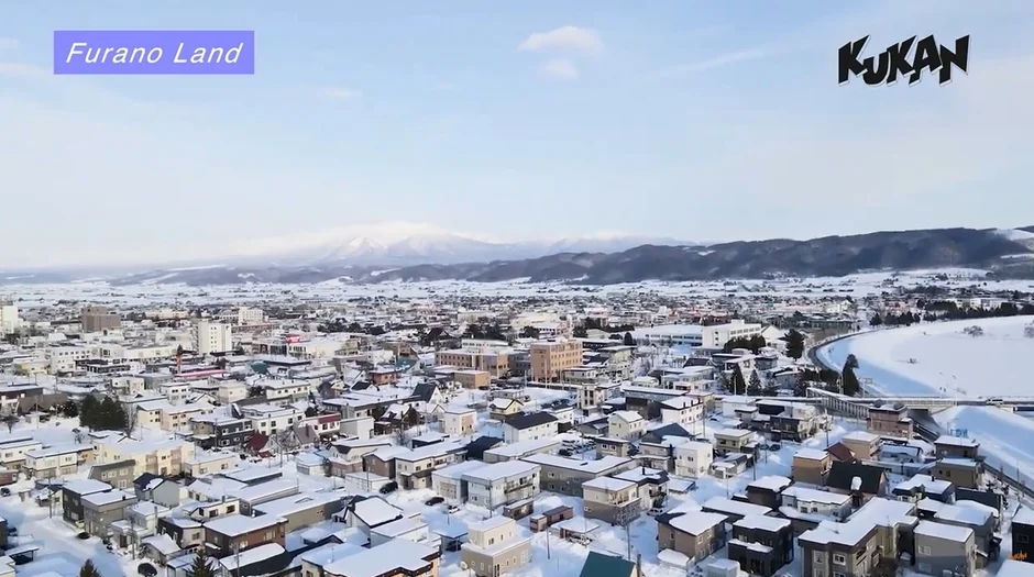 雪に覆われた街並みを空から捉えた冬の風景です。多くの家屋や建物が白く染まり、遠くには雪化粧をした山々が広がっています。富良野の冬の美しい景観を表現しています。