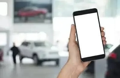 車が並ぶショールームを背景に、白いブランク画面のスマートフォンを片手が持っている様子。モバイルデバイスが自動車販売環境で使用される状況や、画面に情報を表示する前の状態を示唆しています。