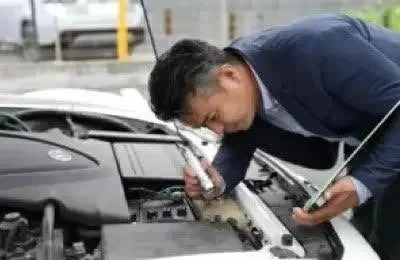 スーツを着た男性が車のボンネットを開け、エンジンルームを覗き込んで点検している様子です。自動車の整備や故障診断を行っている場面と考えられます。